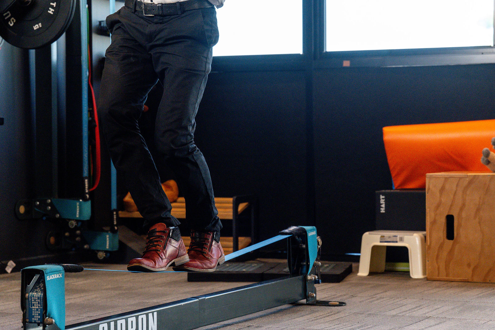 Close-up of a man's feet in leather boots balancing on a Gibbon Slackrack rail, with a HART storage box and orange crash mat in the background