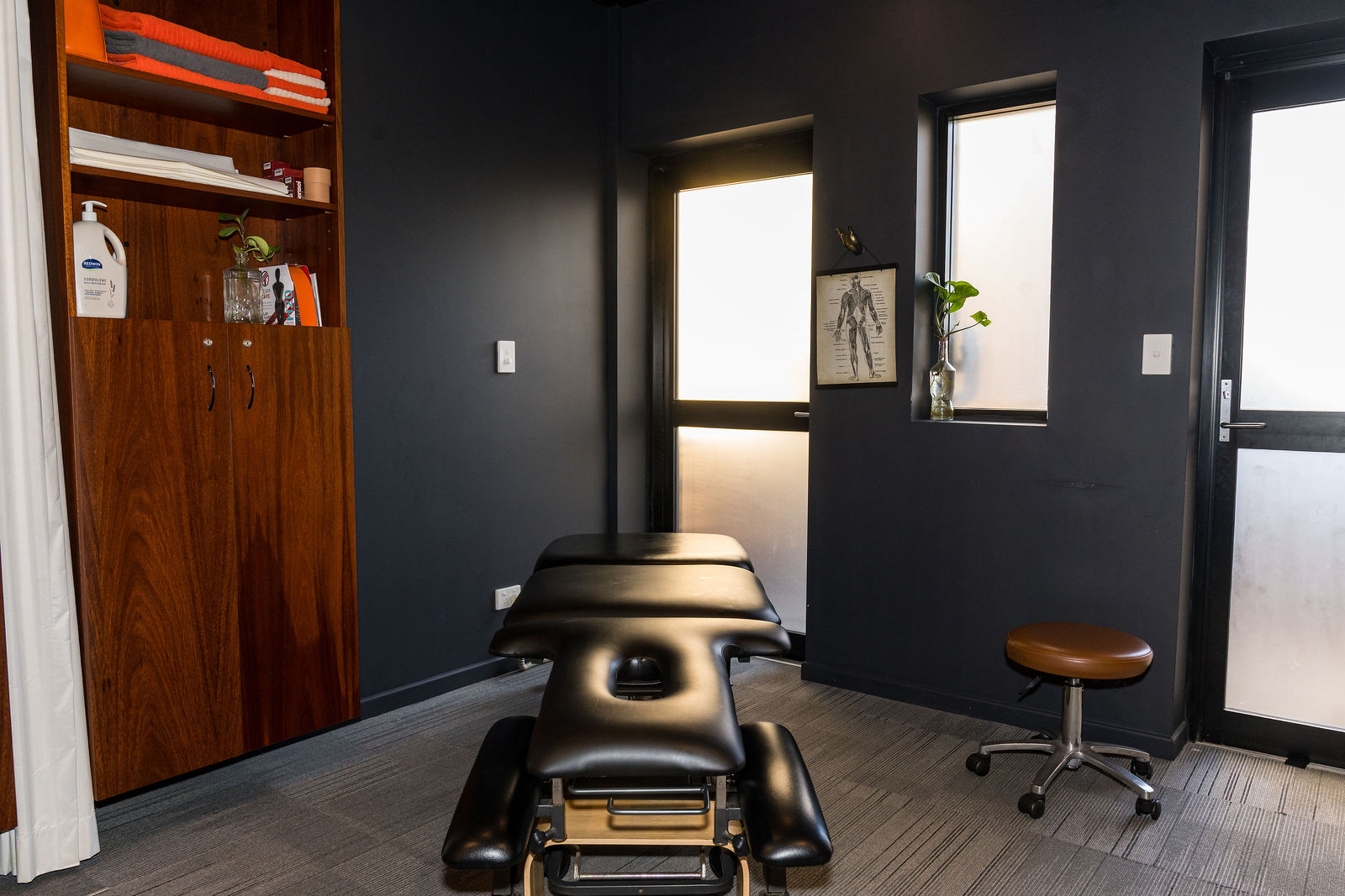 A treatment room at Physio by Design with a plinth, timber storage cabinet, and anatomical chart on the wall