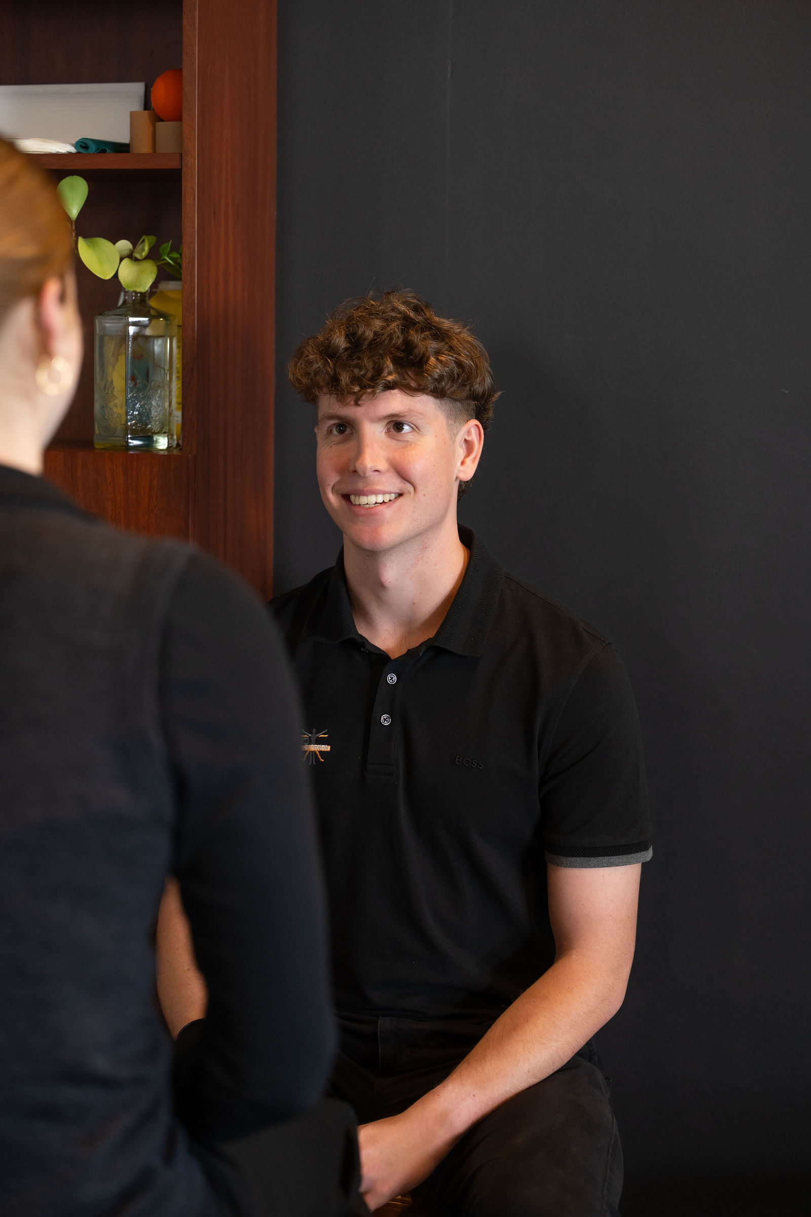 A clinician in a black polo smiling and talking with a client, standing in front of a warm timber shelving unit with plants and treatment props against a dark charcoal wall