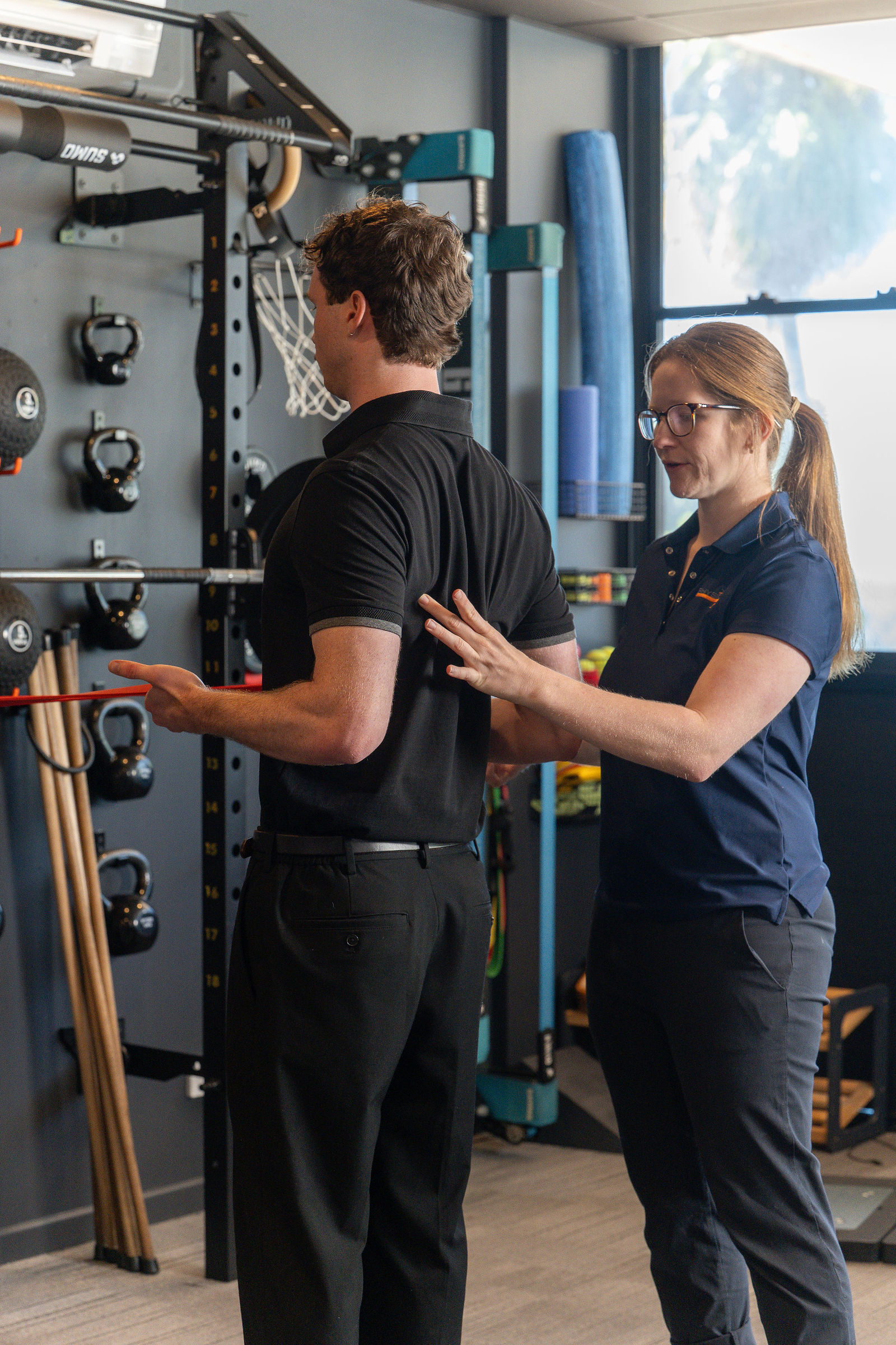 A clinician guiding a client through a resistance band exercise in the gym area, hand on his back for postural feedback, surrounded by kettlebells and a basketball hoop