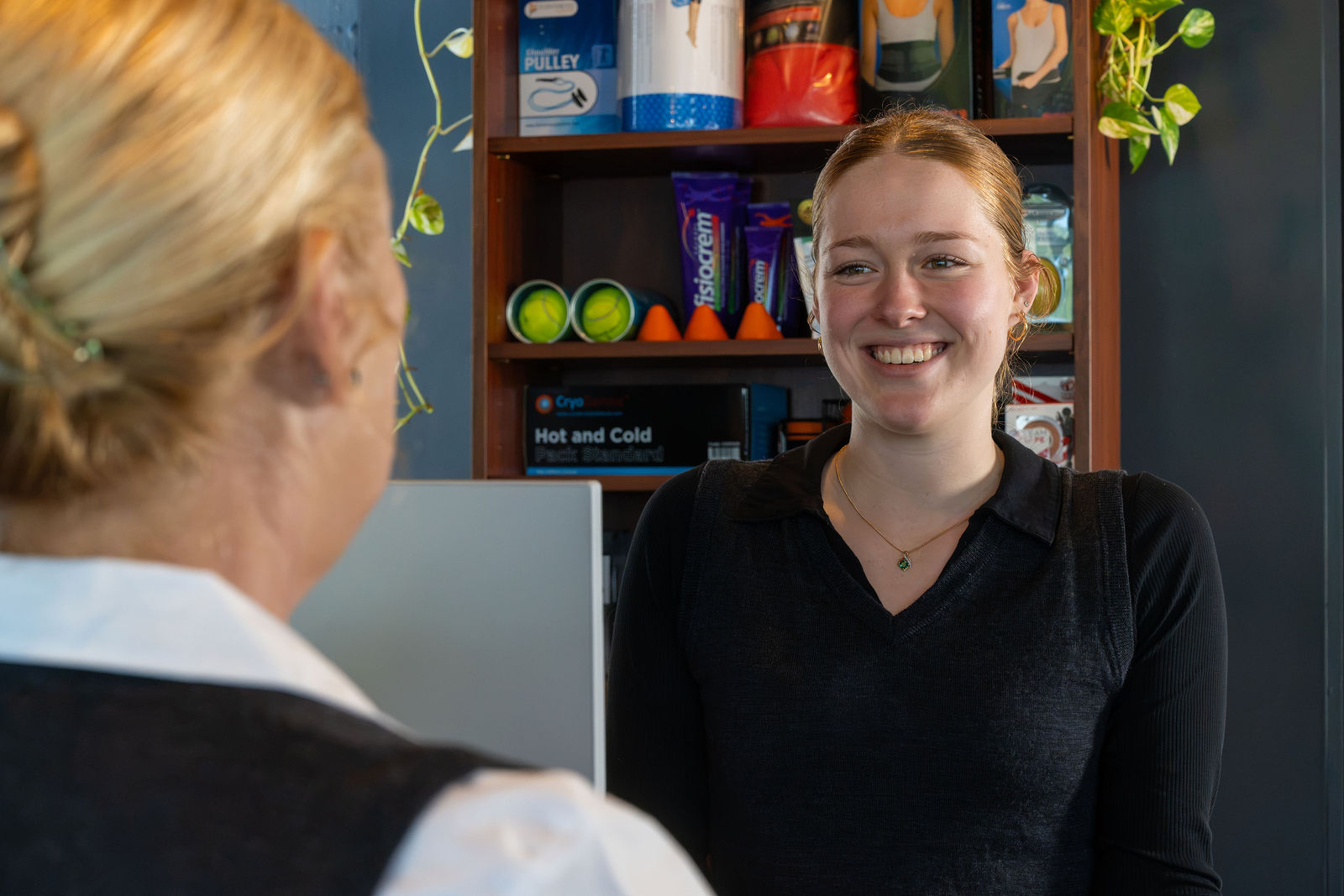 A smiling reception staff member greeting a client at the front desk, with a shelf of physiotherapy retail products and plants in the background