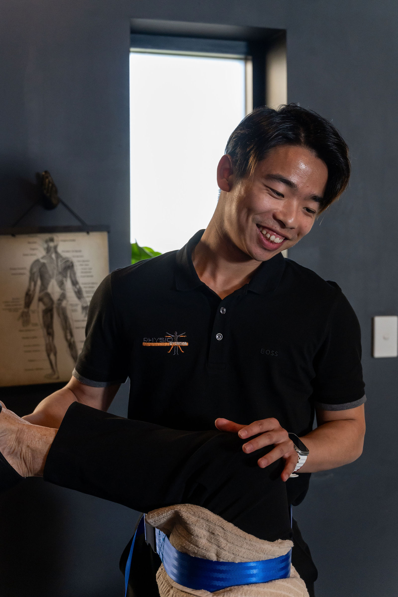 A smiling clinician assessing a client's arm during a hands-on treatment session, with an anatomy chart visible in the background