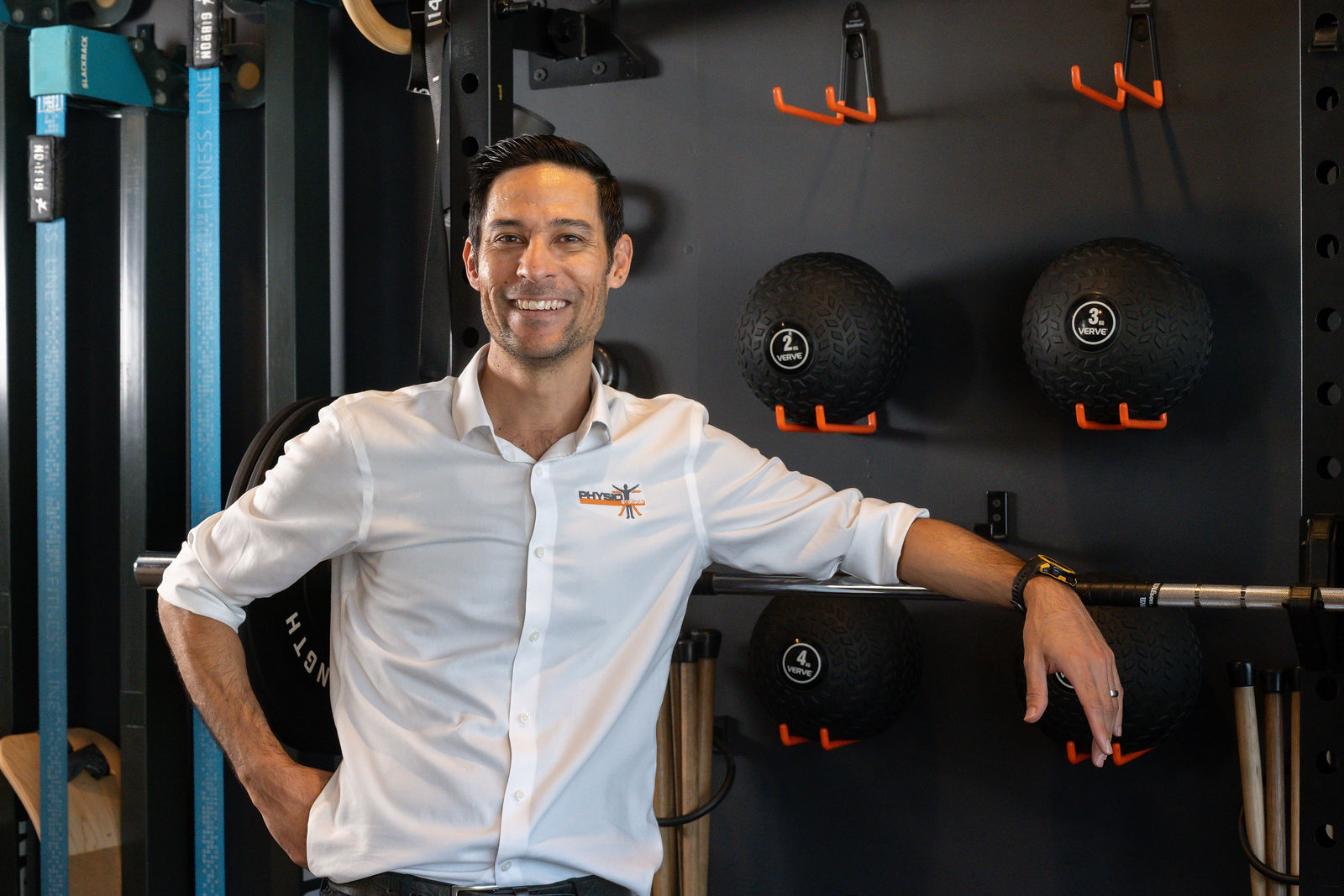 Fabian, Owner & Principal Physiotherapist at Physio by Design, smiling with his hand on his hip in front of a gym equipment wall with slam balls and slacklines