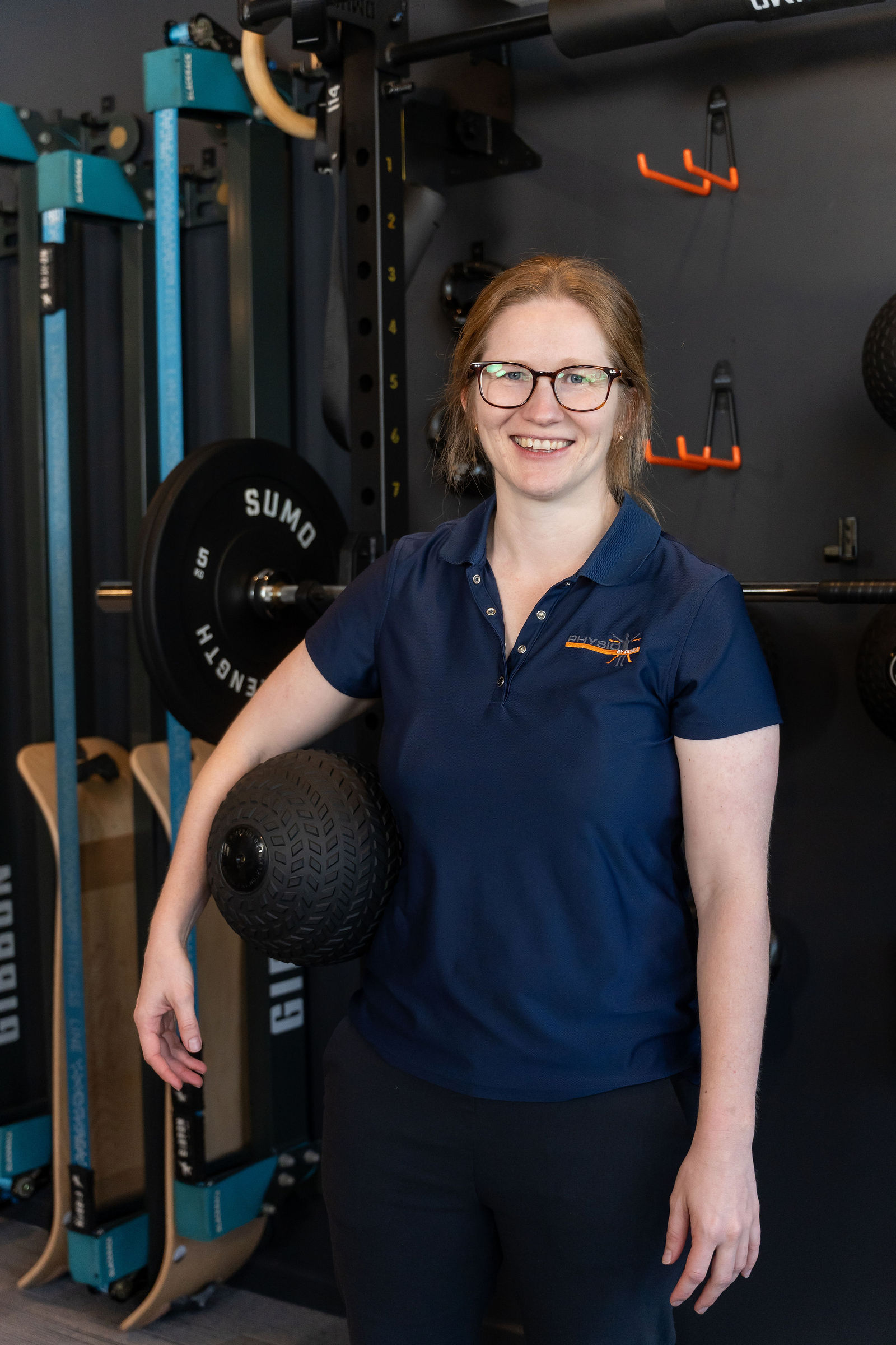 Katie, Exercise Physiologist & Massage Therapist at Physio by Design, smiling with her arm resting on a slam ball in the gym